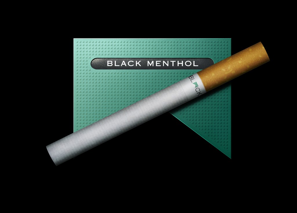 A diagonally-placed cigarette lies atop a black, dark-teal cigarette pack against a black backdrop in a studio product-style shot.