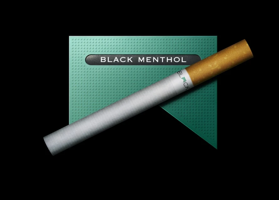 A diagonally-placed cigarette lies atop a black, dark-teal cigarette pack against a black backdrop in a studio product-style shot.