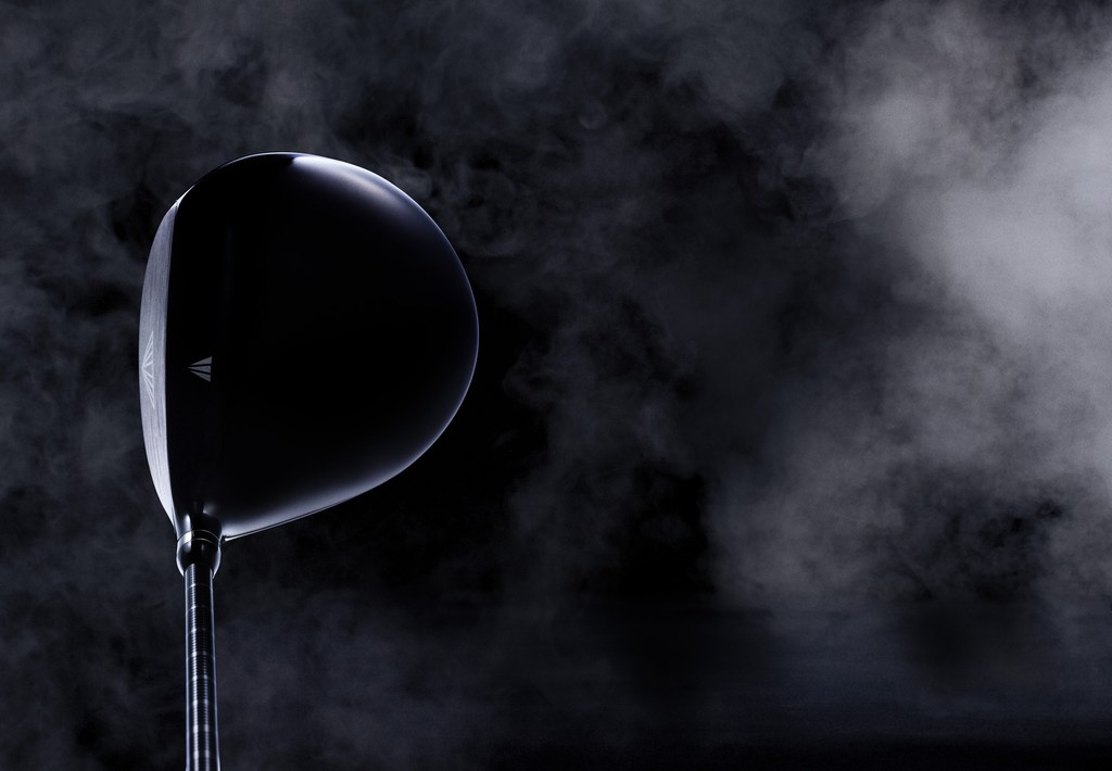 Elevated studio shot showcases a sleek black golf club head against a dark background with smoky gradients, highlighting its modern design.
