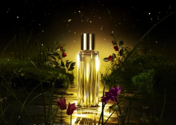 A cylindrical glass perfume bottle with a gold cap standing upright amidst water, mossy rocks, and small colorful flowers under a starry night sky.