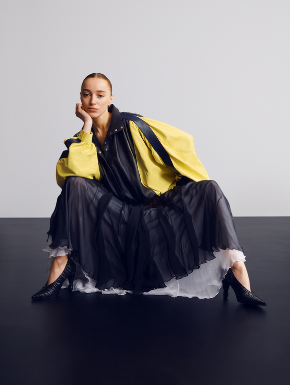 Adult female fashion model poses while sitting in black heels, wearing a black skirt and yellow jacket against a grey and black backdrop.