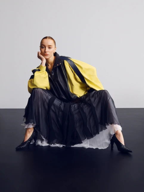 Adult female fashion model poses while sitting in black heels, wearing a black skirt and yellow jacket against a grey and black backdrop.