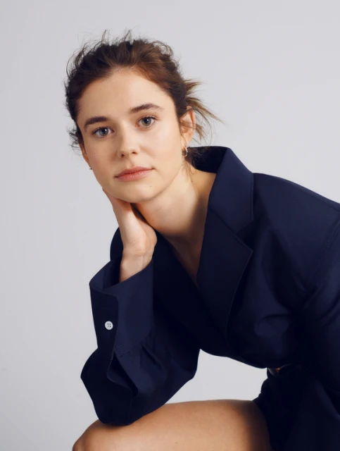 Portrait of a young woman with her hair up, wearing a navy blue jacket, posing in front of a neutral light grey background.