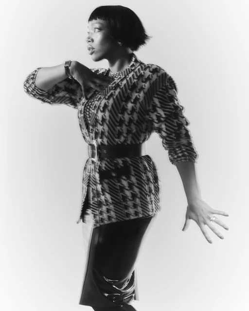 Monochrome portrait of an African-American woman in patterned jacket and black patent skirt, striking a pose against a light background.