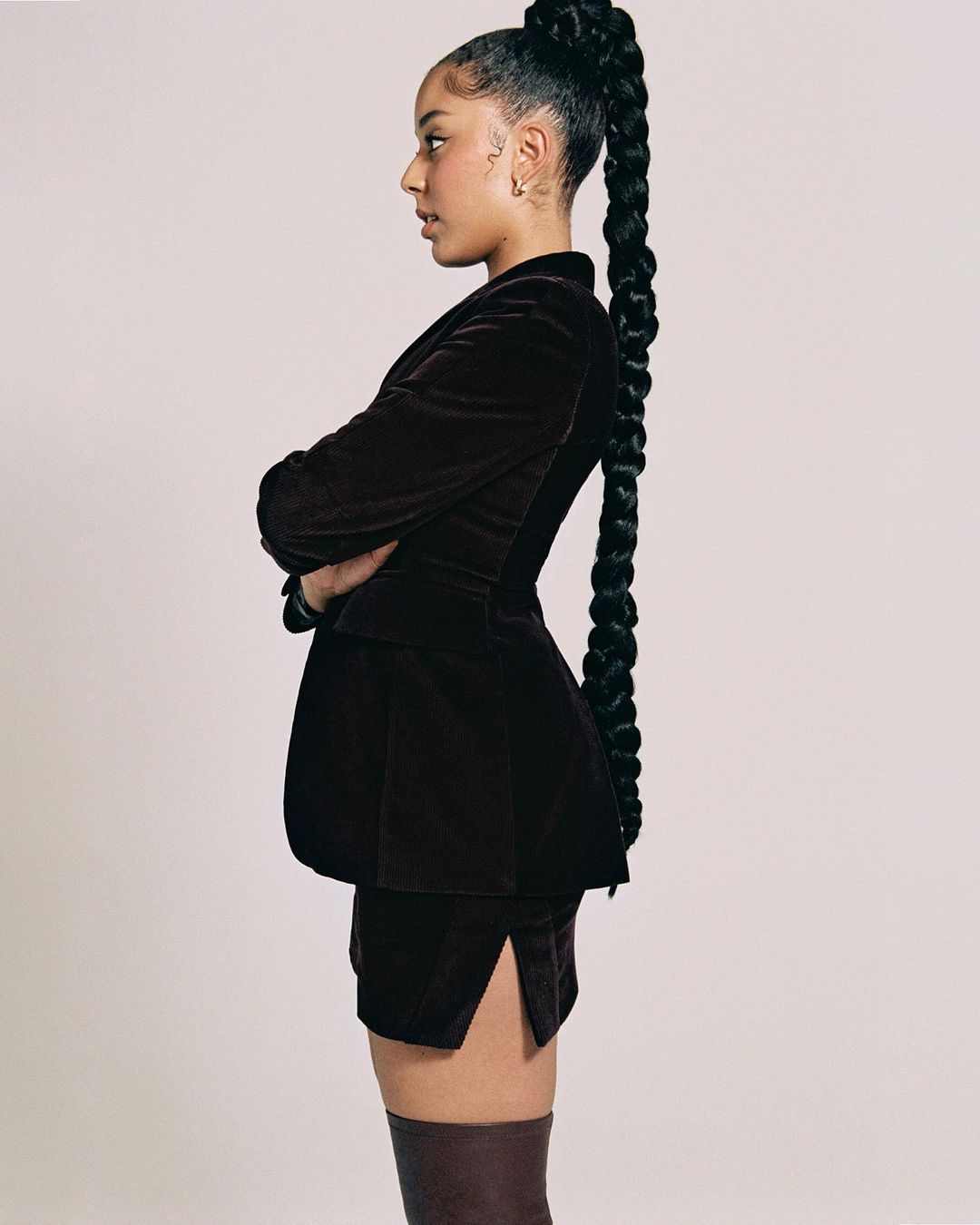 Profile shot of a Black female in a brown velvet skirt suit with her arms crossed, showcasing a long braided ponytail against light gray.