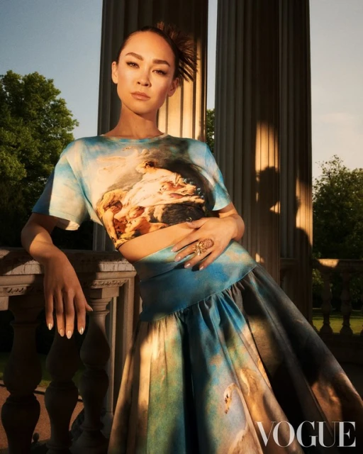 A woman poses near stone columns wearing an art-printed matching skirt and top in an outdoor setting with trees and natural lighting.