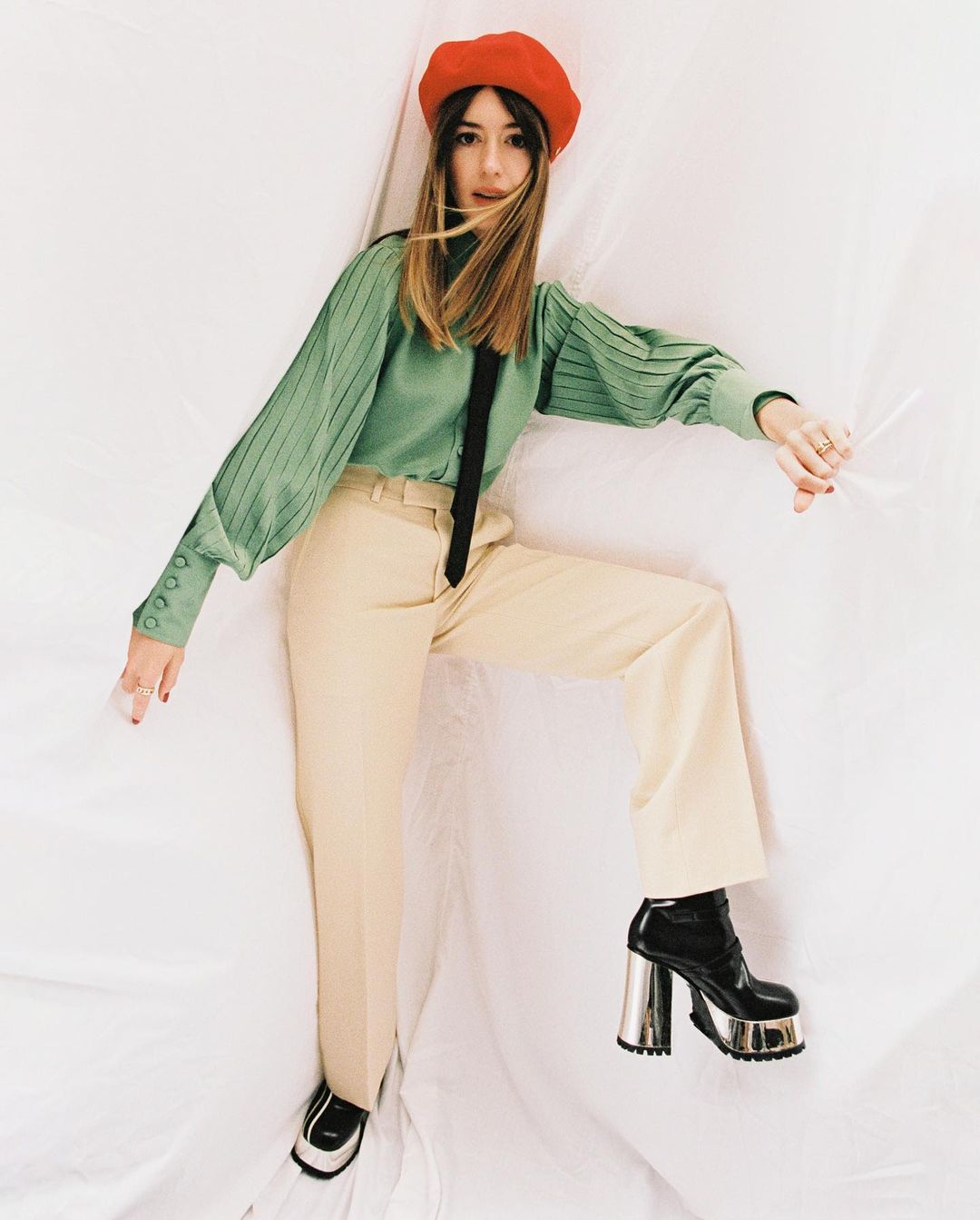Young woman in red beret poses in shiny platform boots, light green top with tie, and beige pants on a white backdrop.