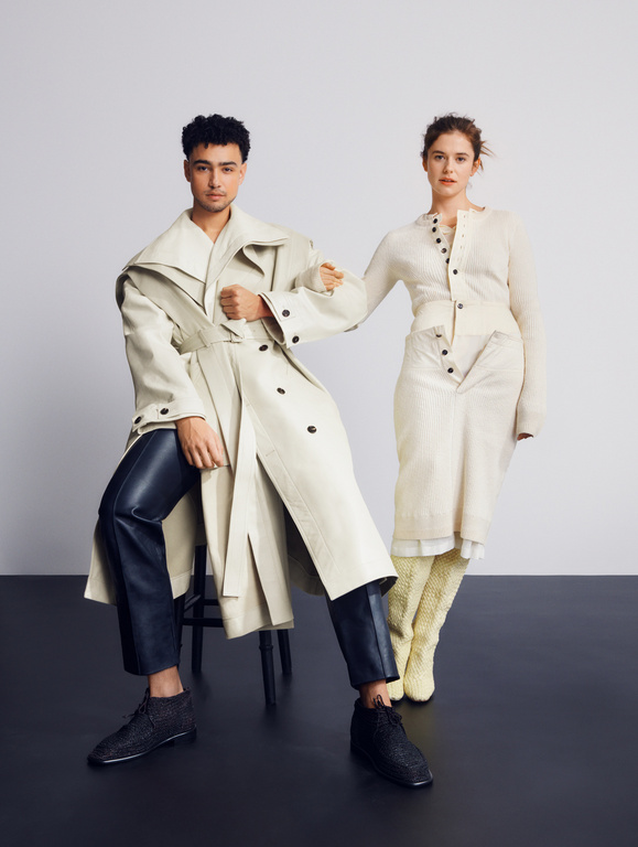 Two fashion models pose in a studio: a man in a trench coat sits on a stool, while a woman in a dress stands beside him.