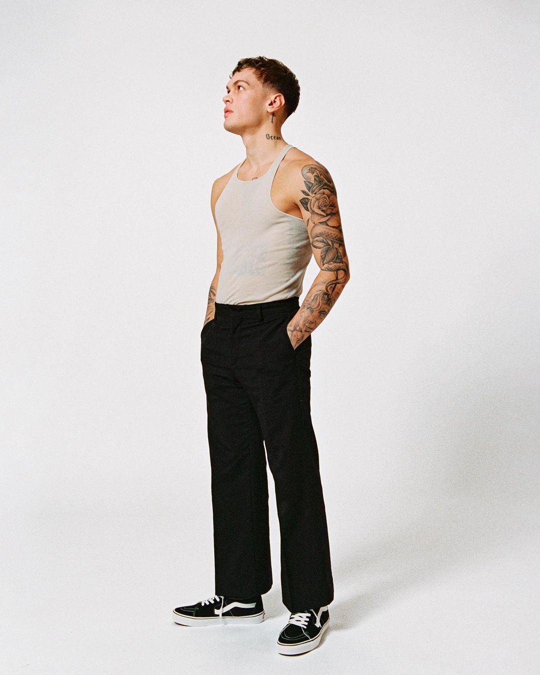 Young man with heavily tattooed arms wearing a cream ribbed tank top and black trousers against a white background.