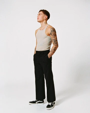 Young man with heavily tattooed arms wearing a cream ribbed tank top and black trousers against a white background.