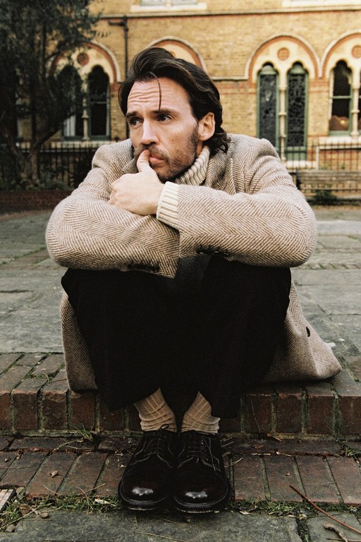 Man sitting on brick steps outside a building, wearing a herringbone coat, dark pants, and black shoes, with an introspective expression.