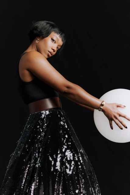 Woman in a black dress poses while holding a white balloon against a dark background in a studio shot.  She is also wearing a wide belt.
