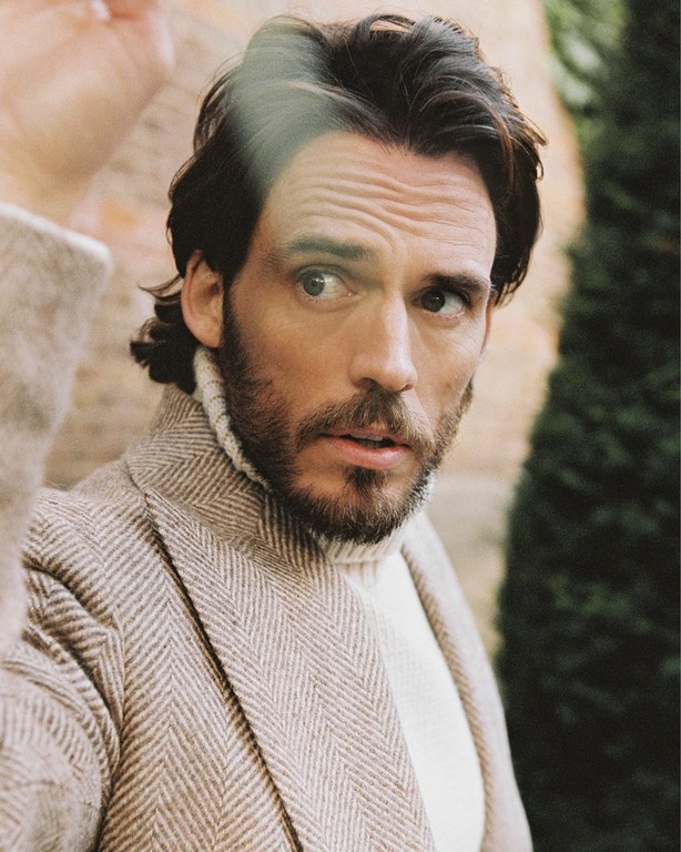 Portrait of a man with brown hair and a beard wearing a beige herringbone jacket and cream turtleneck, gazing sideways in soft light.