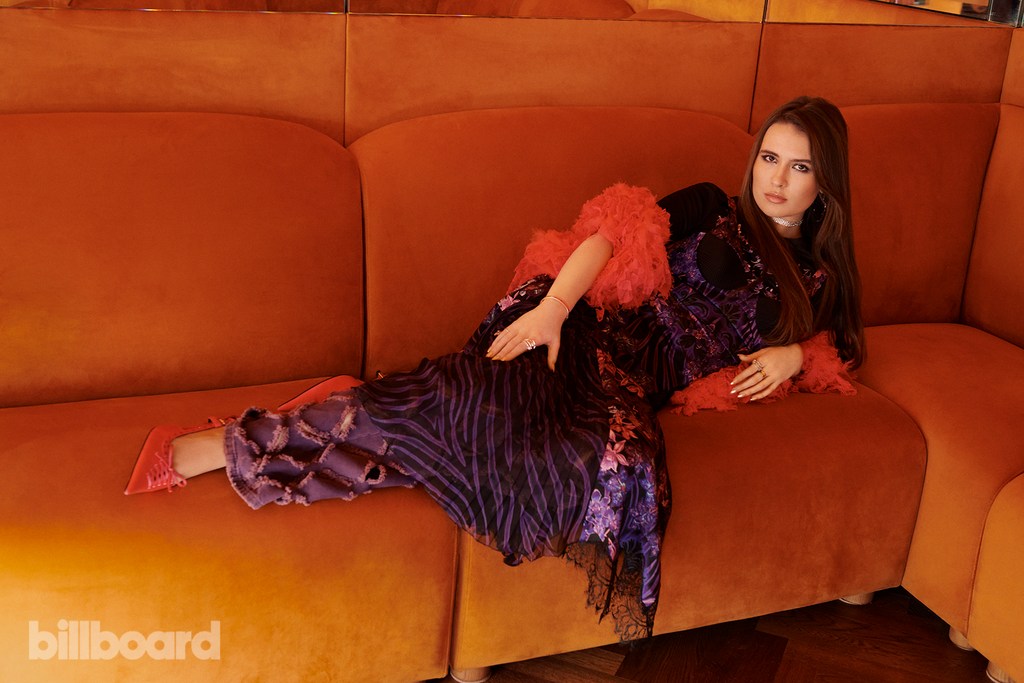 Adult woman in a purple and black floral dress with red feathered trim, lying on an orange couch in an indoor setting.