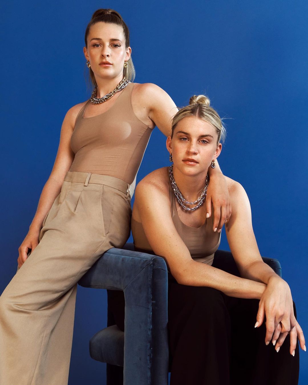 Two women pose in a studio with a blue backdrop, one standing and one seated on a blue chair, wearing tan tops and silver necklaces.