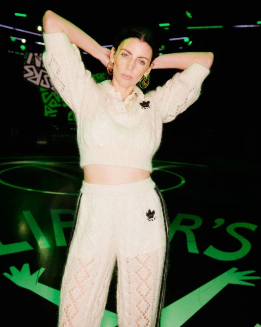 Woman in cream knitwear poses with hands behind head in front of a green stage design, featuring sportswear and athletic apparel fashion.