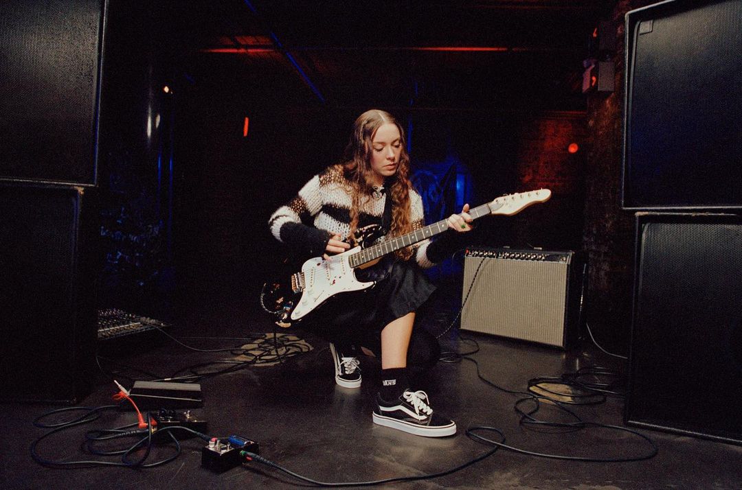 A young adult female playing an electric guitar on a stage with large speakers and audio equipment, captured in a candid, low-lit concert setting.