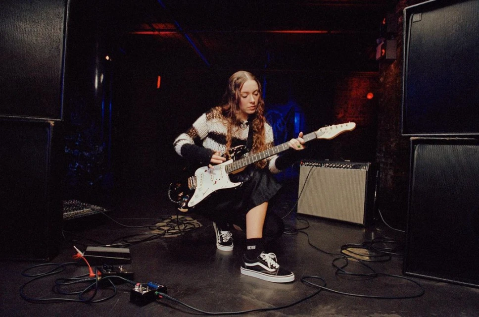 A young adult female playing an electric guitar on a stage with large speakers and audio equipment, captured in a candid, low-lit concert setting.