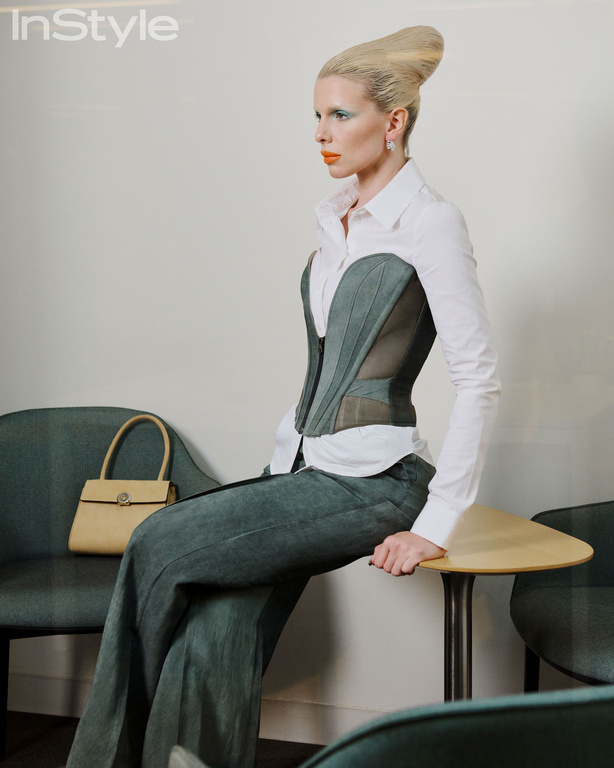 A blonde adult woman in a grey corset, white shirt, and wide-leg pants sits on a yellow-topped table in an indoor setting. Fashion shot.