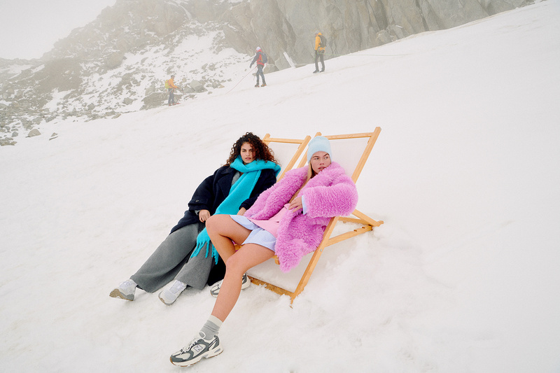 Two women in fashionable winter wear sit on a folding beach chair in a snowy mountain landscape, with hikers in the background.