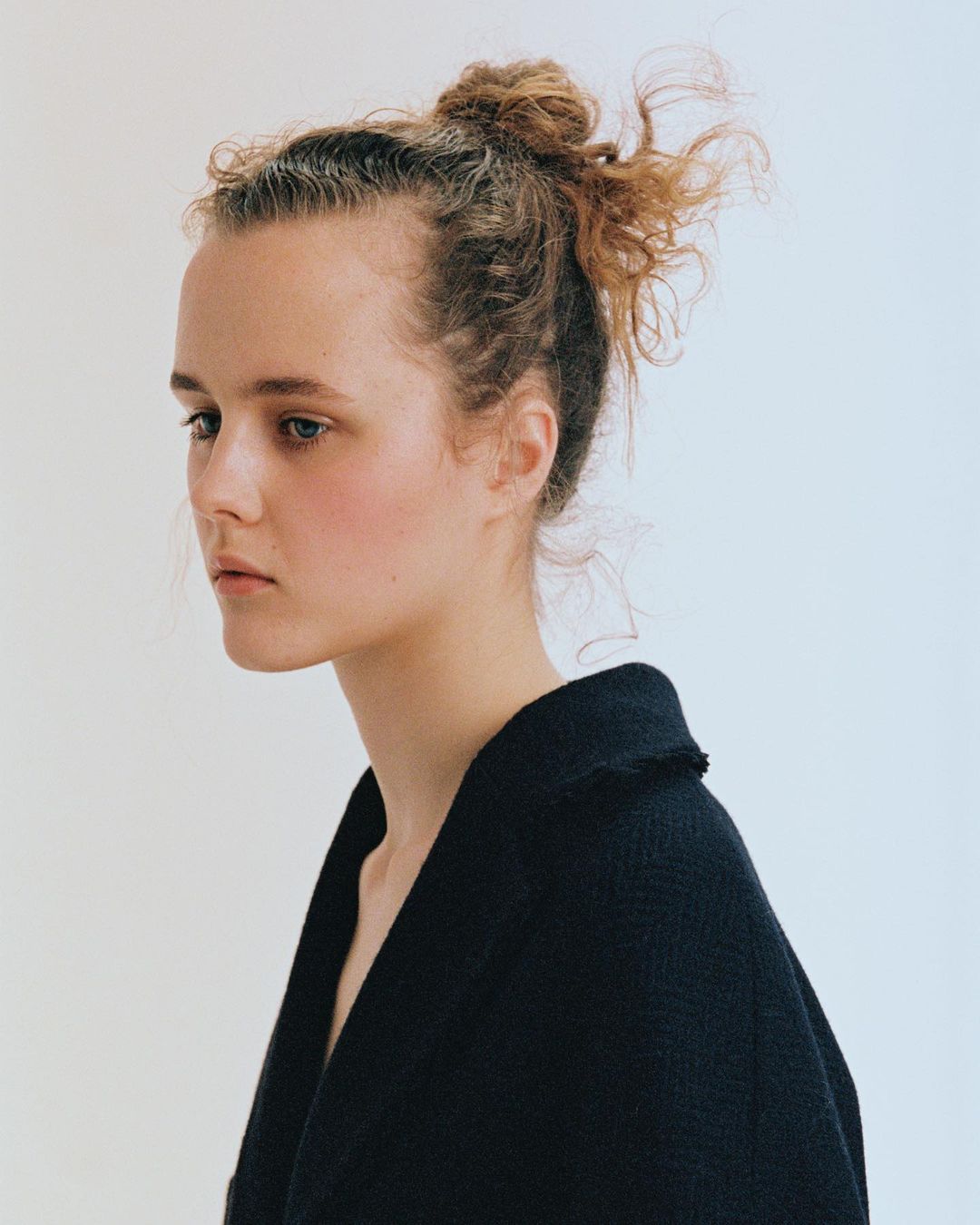 Portrait of a young woman with curly hair in a bun wearing a dark coat against a plain white background in a studio setting.