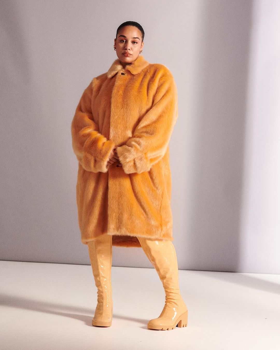 Woman in an orange faux fur coat and high boots poses in a studio against a neutral backdrop, fashion photography.
