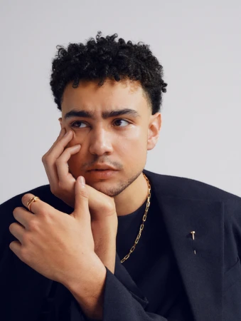 Close-up portrait of a young man with dark curly hair wearing a dark blazer, gold chain necklace, and ring, photographed by Rosaline Shahnavaz.