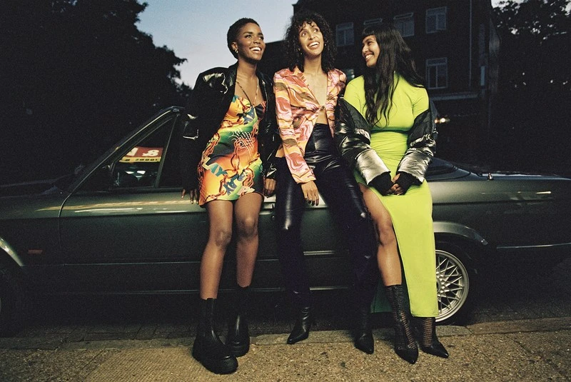 Three women in fashionable outfits pose next to a classic car at dusk, sharing a laugh with colorful dresses and stylish jackets.