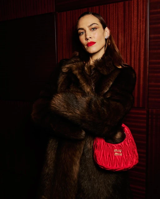 Woman wearing a fur coat and a red purse with the brand logo against a dark red paneled wall in a medium indoor shot.