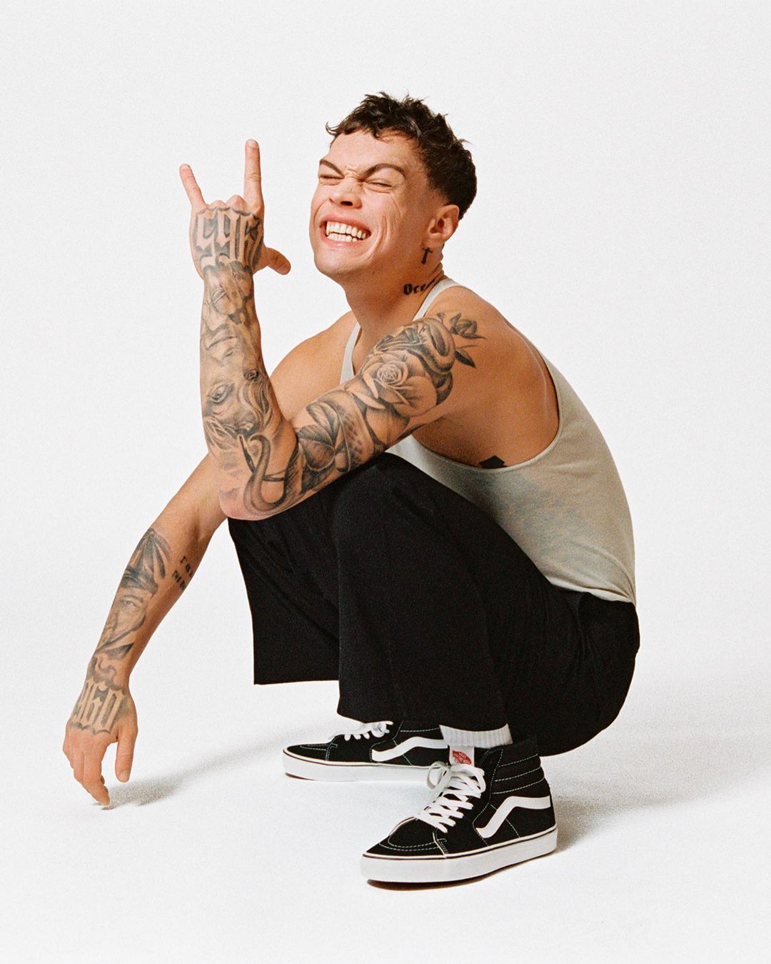 Young adult man in tank top squats in photo studio, smiling with a hand gesture, tattoos, black pants, and Vans sneakers.