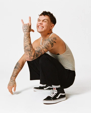 Young adult man in tank top squats in photo studio, smiling with a hand gesture, tattoos, black pants, and Vans sneakers.
