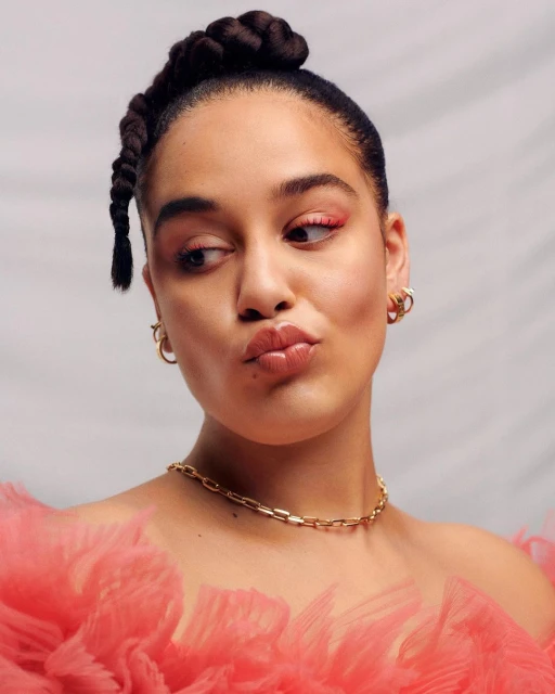Portrait of a woman with braided hair and pink eye makeup, wearing a gold necklace and pink tulle top, against a gray backdrop.