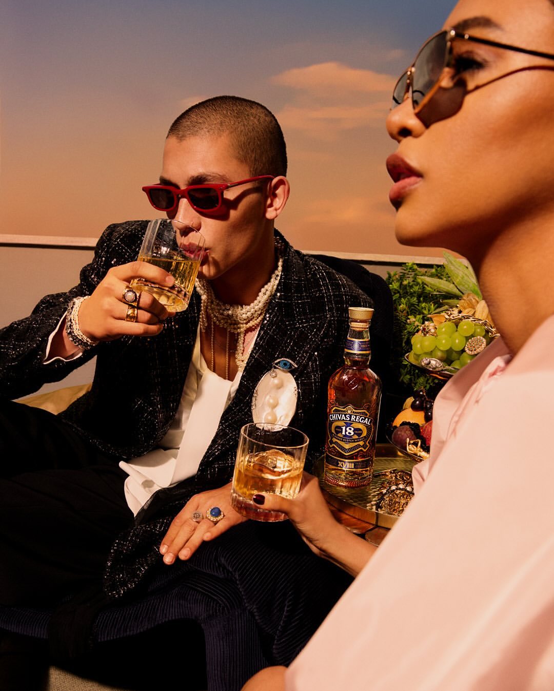 Two stylish people drink whiskey against a golden sky. The woman wears sunglasses and the man has red framed glasses and pearls.