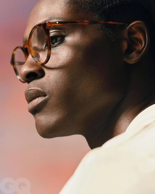 Close-up portrait of a person with dark skin and tortoiseshell glasses, gazing to the side against a soft, gradient background.