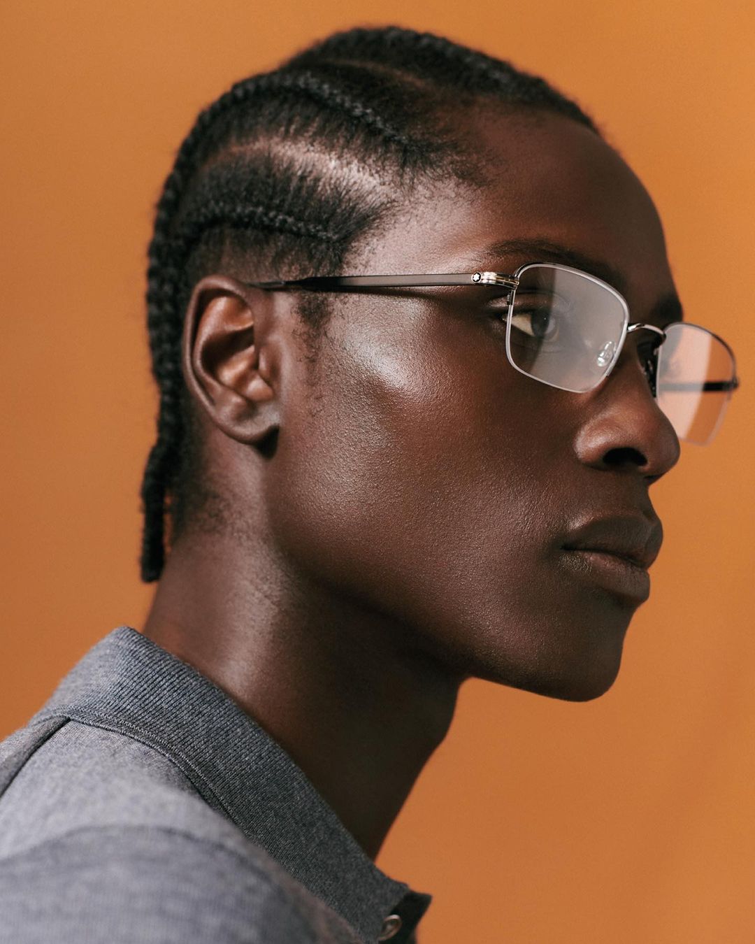 Portrait of dark-skinned man wearing eyeglasses and a gray collared shirt against an orange backdrop in a studio with flattering lighting.