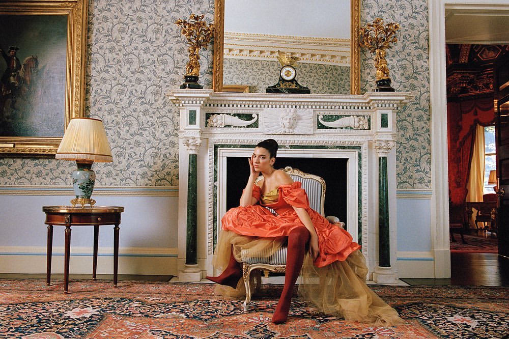 Woman in an orange dress sitting in an ornate chair in a grand room with patterned wallpaper, fireplace, and decorative furnishings.