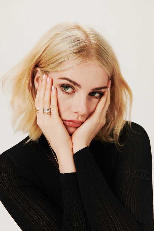 Blonde woman poses in a black long-sleeve shirt against a white backdrop, with her face framed by her hands and fingers.