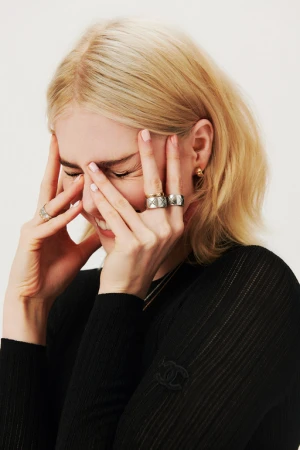 Close-up portrait of a blonde woman wearing rings and a black ribbed top with her hands on her face against a plain background