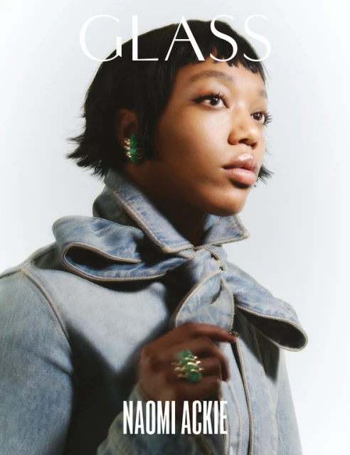 Close-up portrait of Naomi Ackie wearing a denim jacket and green and gold jewellery on a white background for Glass magazine cover.
