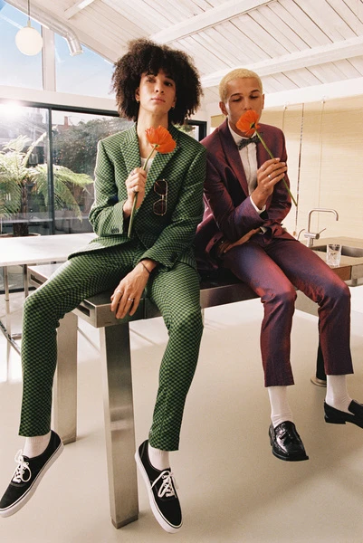 Two young adult male models pose with red flowers in a light-filled studio. One wears a green suit and the other a purple suit.