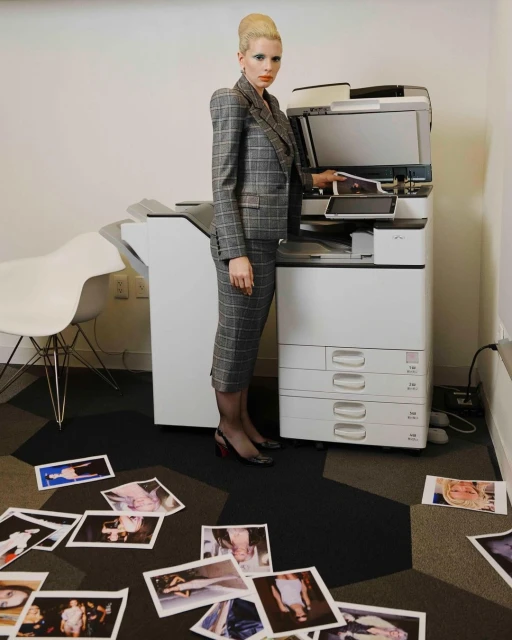 A woman in a grey plaid suit stands at a copier, surrounded by scattered photos, in a brightly lit indoor office environment.