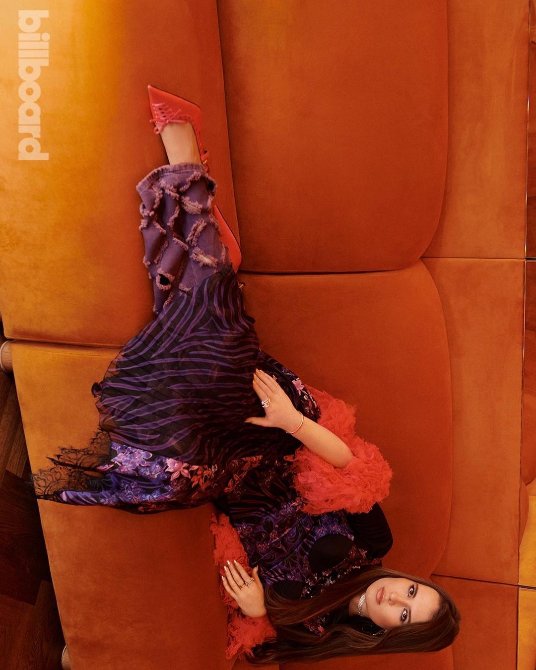A fashionable woman with long brown hair poses on an orange velvet couch, wearing a purple and black gown in a portrait shot.