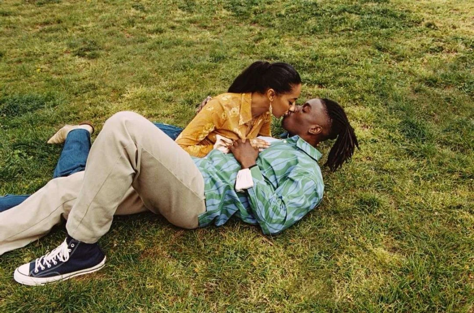 A couple lies together on a grass lawn. The woman is wearing a yellow patterned shirt and the man is wearing a blue and green patterned shirt.
