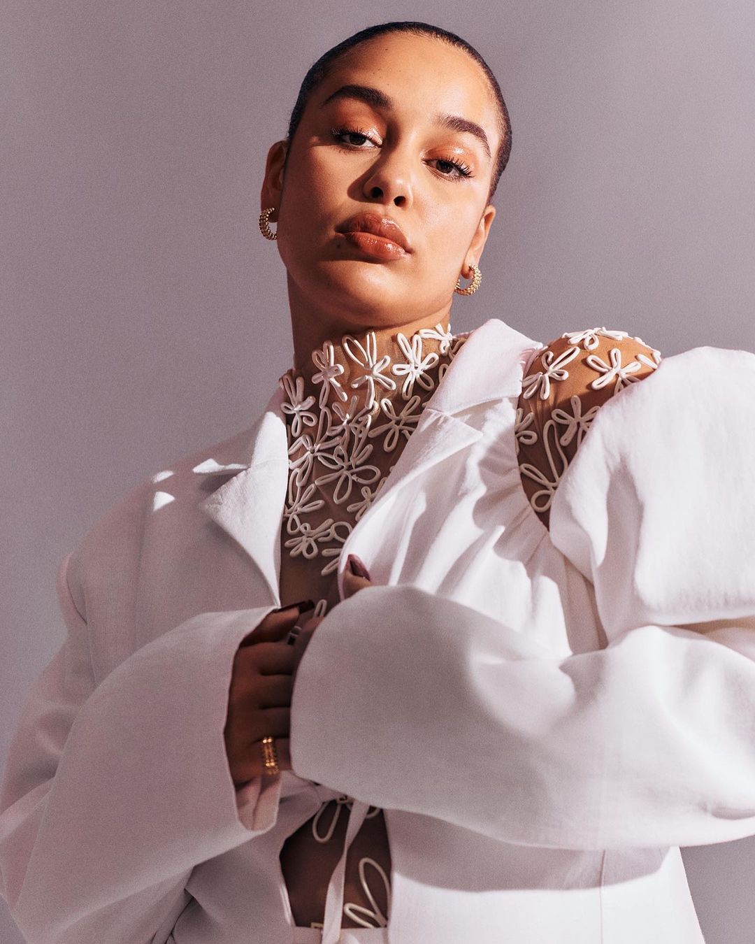 Portrait of a woman wearing a white jacket and a sheer flower-patterned top, posing in front of a solid grey background in a studio setting.