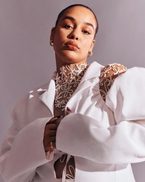 Portrait of a woman wearing a white jacket and a sheer flower-patterned top, posing in front of a solid grey background in a studio setting.
