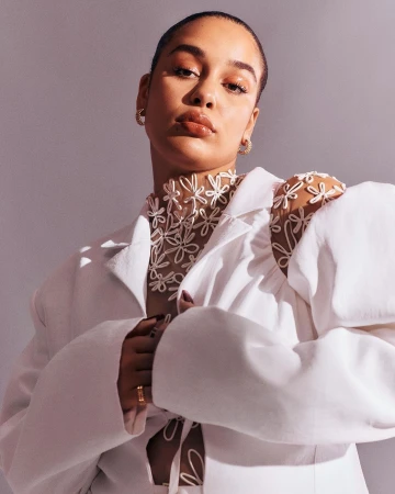 Portrait of a woman wearing a white jacket and a sheer flower-patterned top, posing in front of a solid grey background in a studio setting.