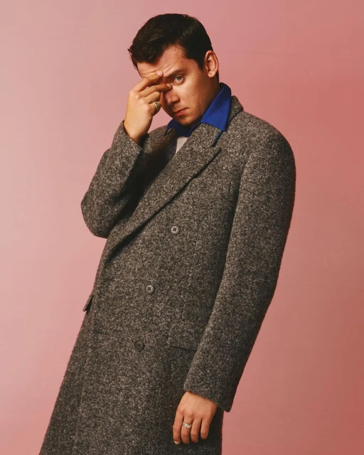 Stylish portrait of an adult male in a grey coat against a pink backdrop, hand to temple, confident gaze, evoking a sense of contemplation.