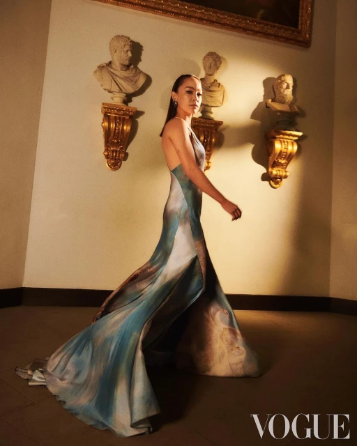 An elegant woman poses indoors in a long watercolor-style dress beside sculptures, with soft lighting for a high-fashion editorial.