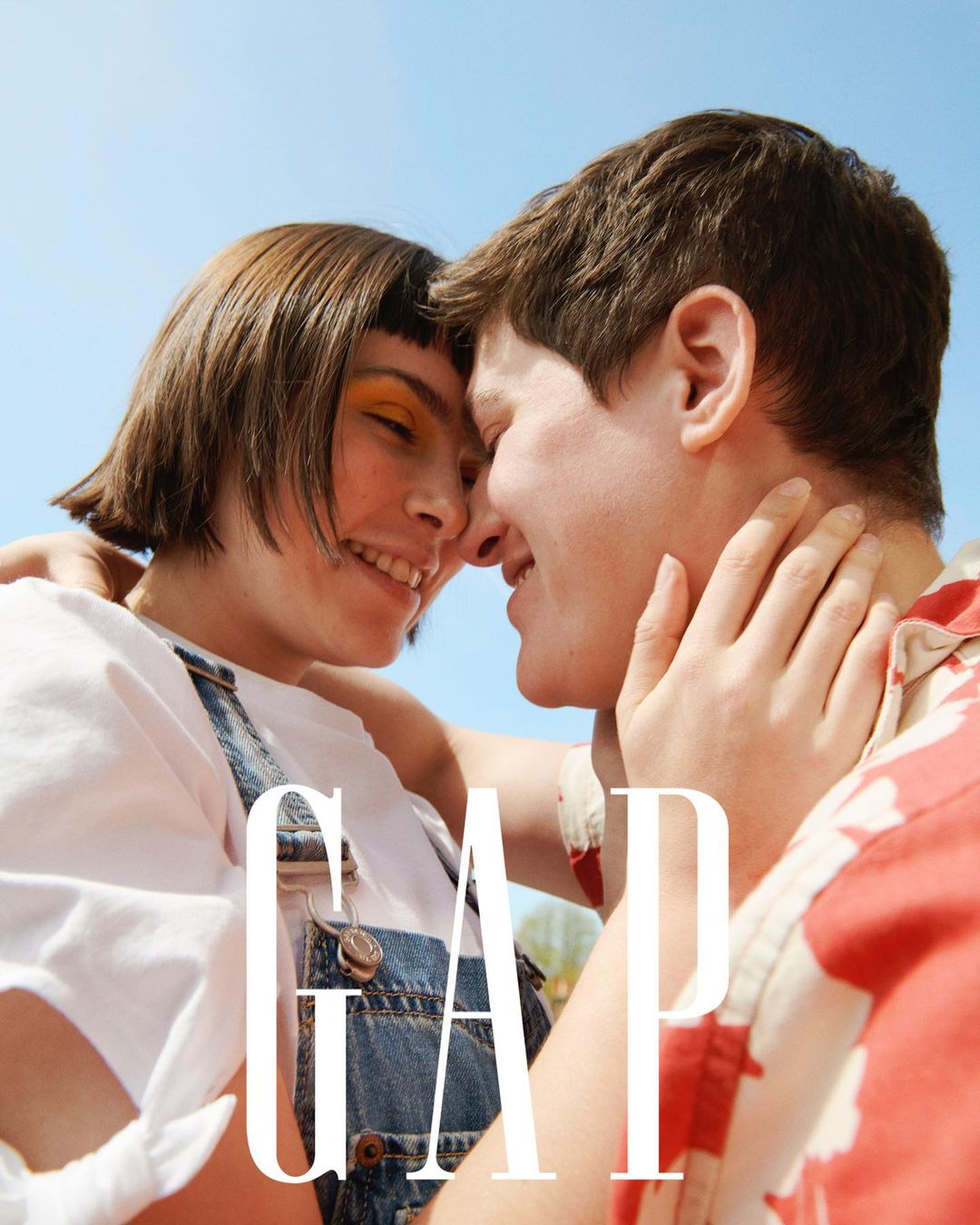Smiling couple embracing outdoors in a close-up shot, wearing a white t-shirt, denim overalls, and a red patterned shirt, with the GAP logo.