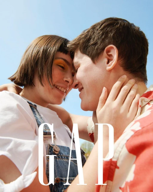 Smiling couple embracing outdoors in a close-up shot, wearing a white t-shirt, denim overalls, and a red patterned shirt, with the GAP logo.